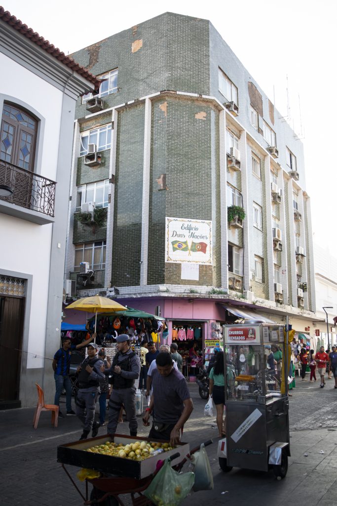 A imagem apresenta a fachada do edifício "Duas Nações", localizado na Rua Grande, um espaço movimentado e característico de comércio popular. O edifício é de esquina, com quatro andares, e sua fachada é revestida por azulejos verdes. As janelas possuem grades ou varandas pequenas com plantas, conferindo um toque de vivacidade ao prédio. Na rua em frente ao edifício, observa-se grande movimento de pessoas e comércio ambulante. Há barracas e carrinhos vendendo frutas e outros produtos, além de guarda-sóis coloridos que protegem os comerciantes do sol. A calçada está cheia de pedestres, reforçando o caráter dinâmico e popular da região.