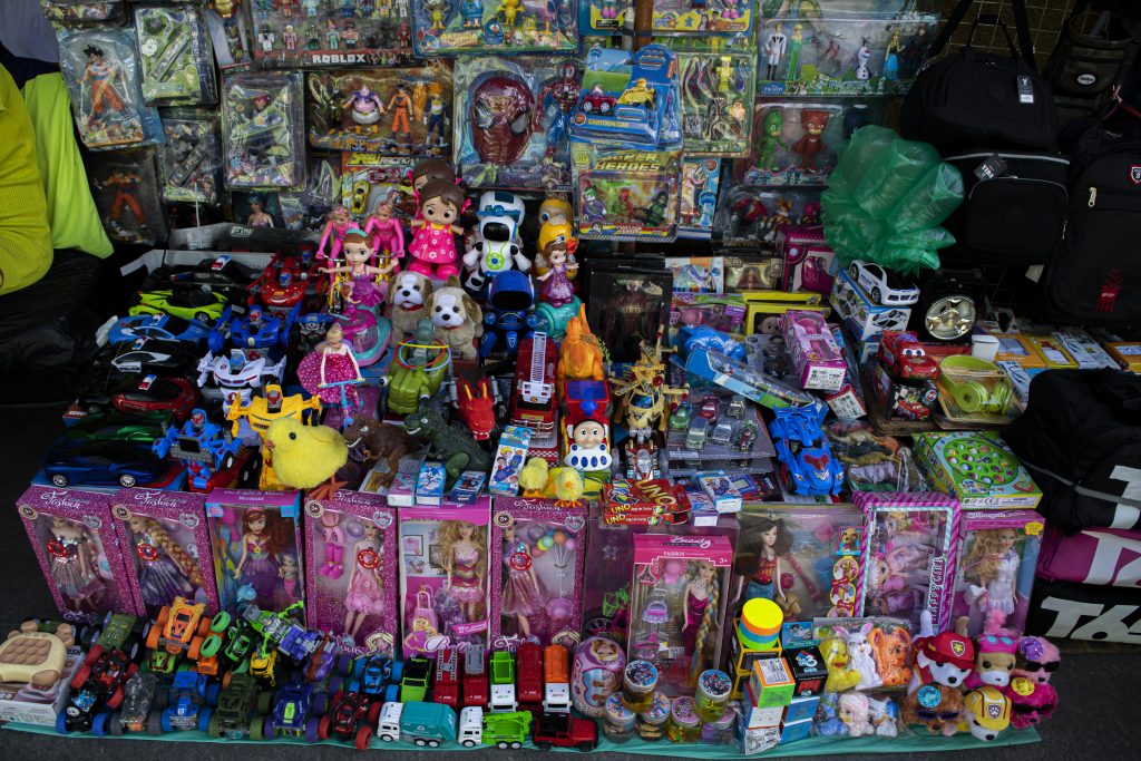Em primeiro plano, sobre uma superfície plana, há uma grande variedade de brinquedos organizados lado a lado. Entre eles, destacam-se bonecas em caixas cor-de-rosa, carros em miniatura, pelúcias de animais, figuras de ação e outros itens infantis. Alguns brinquedos estão soltos, enquanto outros permanecem em suas embalagens originais, exibindo cores vibrantes e ilustrações chamativas. Ao fundo, presos em um painel ou dispostos verticalmente, veem-se mais brinquedos em embalagens plásticas, incluindo figuras de ação de diferentes personagens e temas. À direita, há mochilas ou bolsas pretas empilhadas, reforçando o aspecto de um local de venda informal. Pessoas caminham ao fundo, embora estejam fora de foco, sugerindo uma área comercial movimentada.
