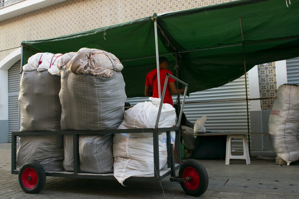 Cena de rua retratando um carrinho de carga metálico com rodas vermelhas, carregado com grandes sacos volumosos de ráfia, amarrados e repletos de mercadorias. Os sacos ocupam todo o espaço do carrinho e estão dispostos em pé, comprimidos uns contra os outros. Ao fundo, um homem de camiseta vermelha, de costas, está parcialmente coberto por uma lona verde presa em estrutura metálica típica cobertura de barraca de feira. O ambiente sugere o início de montagem de uma banca de comércio ambulante em frente a uma loja fechada com porta de metal enrolável. No canto direito da imagem, outro grande saco está no chão ao lado da barraca, enquanto uma mesa simples com uma sacola plástica e um banquinho branco completam a cena. O piso é de paralelepípedos e o prédio ao fundo tem azulejos decorativos azul e branco.