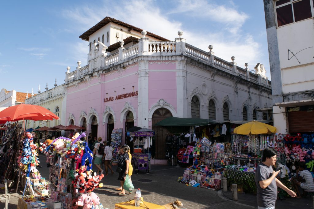 A imagem apresenta uma cena típica do comércio da Rua Grande, em São Luís, Maranhão. No centro, destaca-se um prédio histórico pintado de rosa claro com detalhes brancos, que abriga atualmente uma unidade das "Lojas Americanas". Este edifício possui uma arquitetura marcante, com características coloniais, como arcos nas janelas e portas, além de ornamentos decorativos na fachada. No topo, há um pequeno torreão e uma balaustrada branca que reforçam o estilo clássico do prédio. Historicamente, este edifício abrigava um cinema, evidenciando a transformação de construções históricas em estabelecimentos comerciais ao longo do tempo. Em frente ao prédio, observa-se um fluxo considerável de pessoas caminhando pela rua, caracterizando o movimento intenso típico da área comercial. Além disso, há diversas barracas e vendedores ambulantes espalhados pela calçada. As barracas são cobertas por guarda-sóis coloridos, como vermelho e amarelo, e oferecem uma ampla variedade de produtos, incluindo brinquedos, roupas e acessórios. A cena reflete a convivência entre o patrimônio arquitetônico e a dinâmica do comércio popular que caracteriza a Rua Grande.