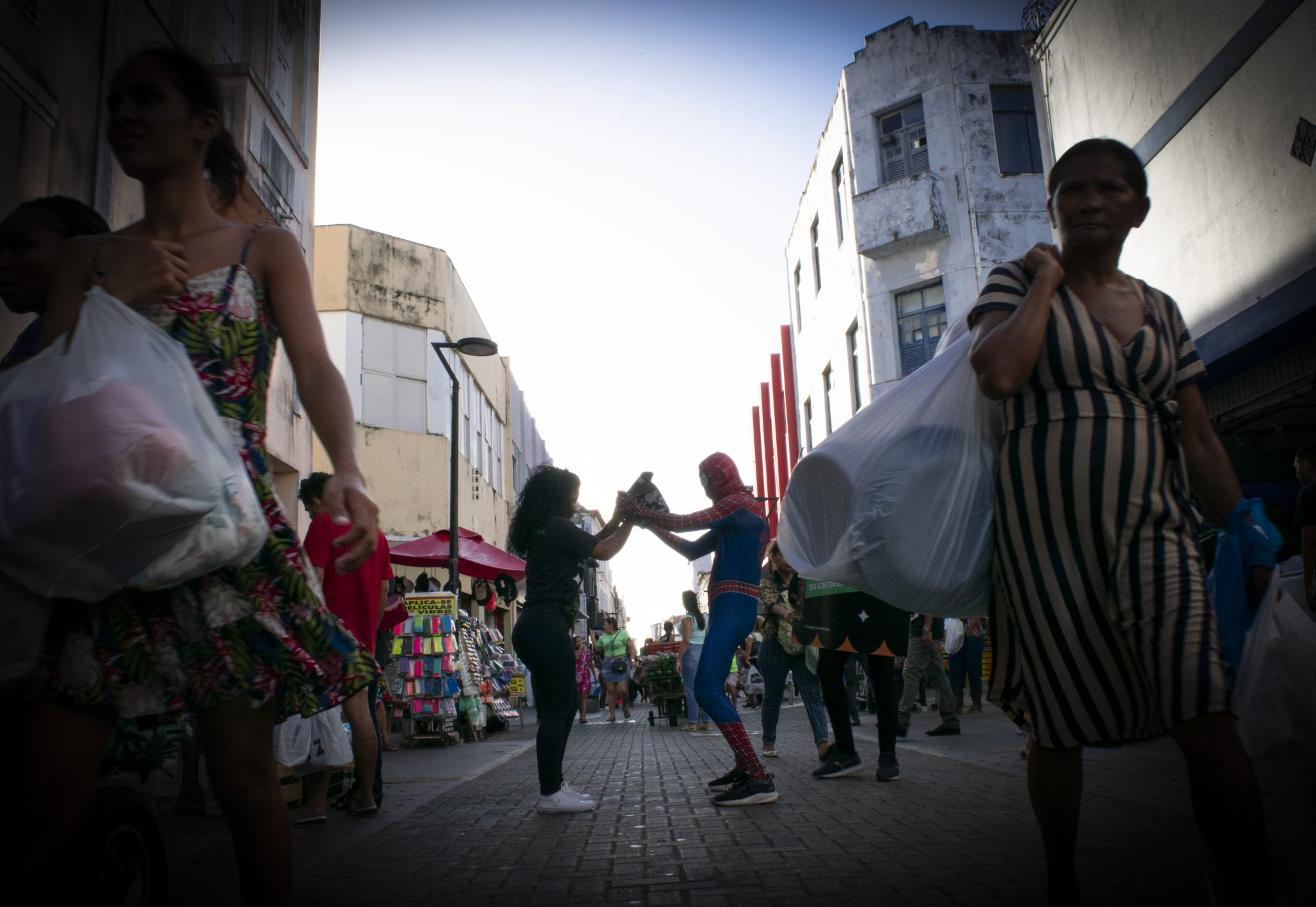 A imagem retrata uma cena movimentada na Rua Grande, um espaço urbano caracterizado por sua arquitetura de prédios antigos e comerciais. As construções ao fundo possuem fachadas desgastadas, com pintura descascada e janelas simples, remetendo a um estilo arquitetônico tradicional e funcional. À direita, há um edifício de cor clara com detalhes em azul escuro, enquanto à esquerda, outro prédio exibe tons amarelados e sinais de desgaste. No centro da imagem, dois vendedores chamam a atenção. Um deles está vestido como o Homem-Aranha, usando um traje azul e vermelho característico do personagem, enquanto o outro está de roupa casual. Juntos, eles estendem as mãos para formar um arco, criando uma interação lúdica no meio da rua. Ao redor deles, várias pessoas caminham carregando sacolas grandes, sugerindo um ambiente comercial ativo. A cena é emoldurada por um efeito de vinheta que escurece as bordas da imagem, diminuindo a visibilidade dos rostos das pessoas nas laterais. Nas margens da rua, barracas coloridas e produtos expostos reforçam o clima de comércio da Rua Grande. A iluminação natural destaca o centro da imagem enquanto as bordas permanecem mais sombreadas, criando um contraste que direciona o olhar para a interação central.
