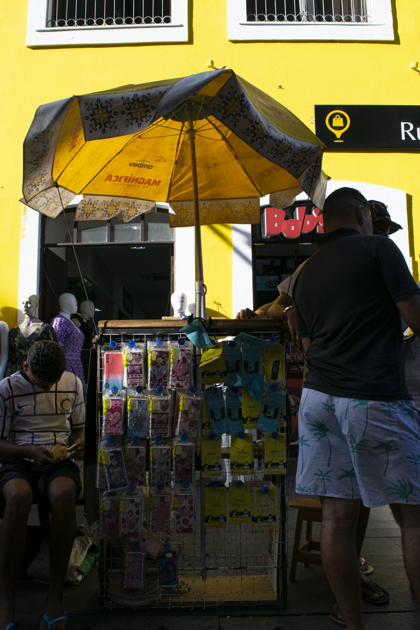 A imagem retrata uma cena típica de comércio informal na Rua Grande. Em primeiro plano, há uma barraca de camelô coberta por um guarda-sol amarelo com detalhes que remetem aos azulejos de São Luís, que oferece sombra para os produtos expostos. A estrutura da barraca é composta por uma grade metálica onde estão pendurados diversos pacotes coloridos, aparentemente contendo capas de celular e acessórios similares. Alguns itens estão organizados em embalagens plásticas transparentes, e há panos ou tecidos dobrados pendurados em um dos lados. À esquerda da imagem, um homem está sentado em um banco. Ele veste uma camiseta listrada branca com detalhes em azul escuro e claro, além de um short preto. Ele segura um celular em uma das mãos, aparentando estar entretido com o aparelho. No lado direito, outro homem está de pé próximo à barraca. Ele veste uma camiseta preta e um short branco estampado com desenhos de coqueiros verdes. Ao fundo, a arquitetura é característica de construções históricas. O prédio tem uma fachada amarela vibrante com janelas brancas e grades pretas nas partes superiores. Há também placas comerciais visíveis, como a de uma loja com o nome "Bob's" parcialmente exibido e outra placa preta com um ícone de lâmpada amarela, indicando possivelmente outro estabelecimento comercial. Manequins vestidos com roupas coloridas podem ser vistos dentro da loja ao fundo, sugerindo a venda de vestuário no local.