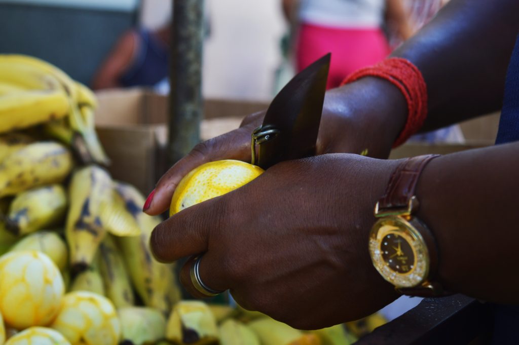 Uma pessoa de pele negra descasca uma laranja utilizando uma faca. Ela usa um relógio dourado no pulso esquerdo e uma pulseira vermelha no pulso direito. No fundo, há caixas com bananas na Rua Grande.