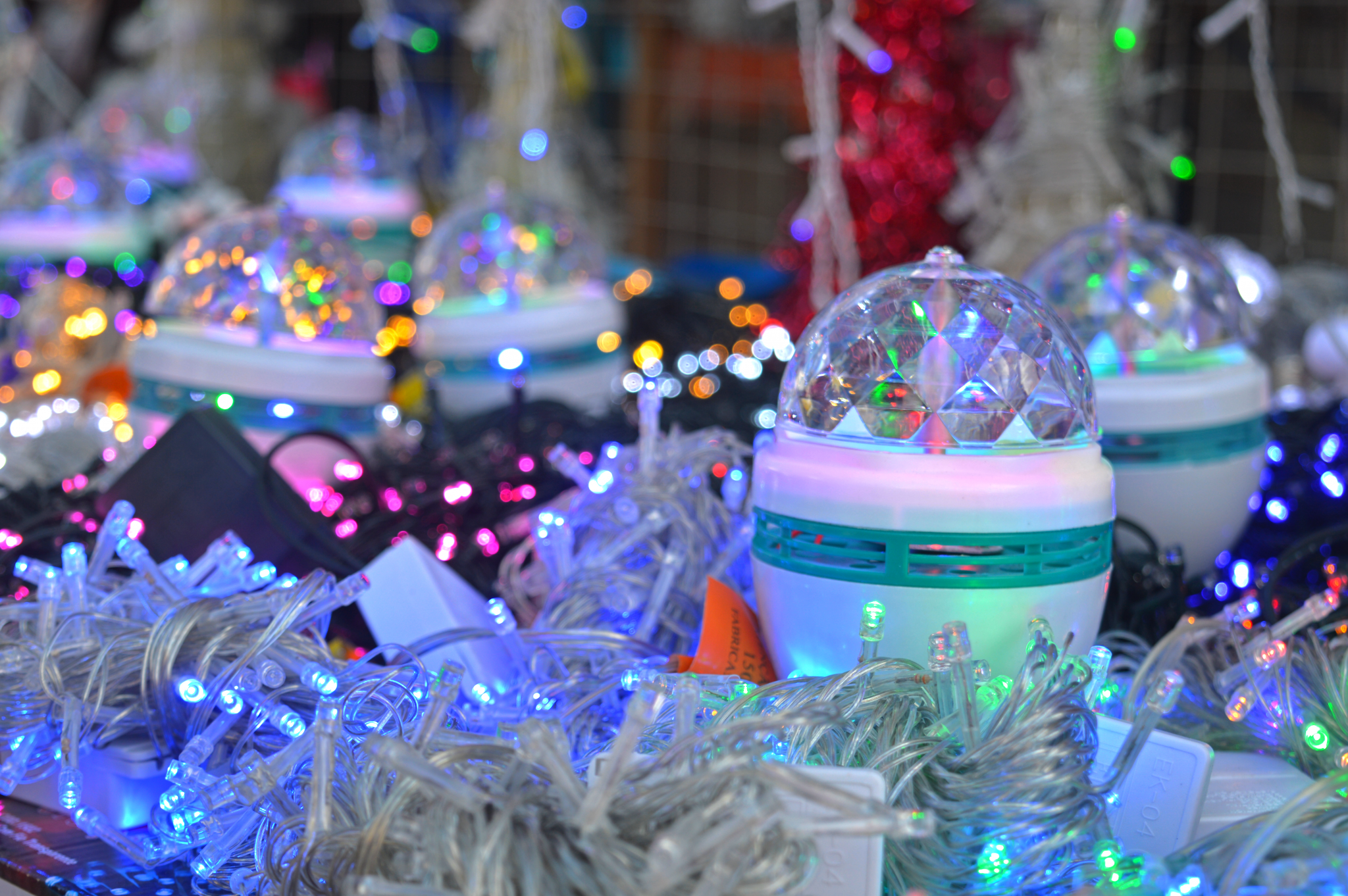 A imagem mostra diversos itens de decoração iluminados, incluindo luzes coloridas e pequenos globos com design facetado que refletem as cores das luzes ao redor. Os globos têm uma base branca com detalhes em verde e estão cercados por fios de luzes LED em tons variados, como azul, rosa e branco.