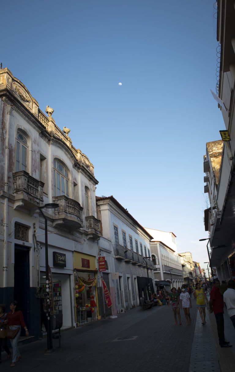 A imagem retrata uma cena urbana da Rua Grande, em São Luís do Maranhão, durante o entardecer. O céu azul domina a parte superior da fotografia, com a Lua visível ao fundo, desfocada e distante, adicionando um toque celestial à composição. À esquerda, destacam-se edificações históricas de estilo colonial, com fachadas ornamentadas e varandas de ferro trabalhado. As janelas altas e os detalhes arquitetônicos remetem ao período de influência portuguesa na região. Do lado direito, há construções mais simples, mas ainda preservando traços históricos. A rua está movimentada, com pessoas caminhando em ambas as direções. Algumas estão próximas às lojas que ocupam o térreo dos edifícios, enquanto outras seguem em pequenos grupos. As vitrines exibem produtos variados, incluindo roupas e acessórios, sugerindo um espaço comercial ativo e popular.
