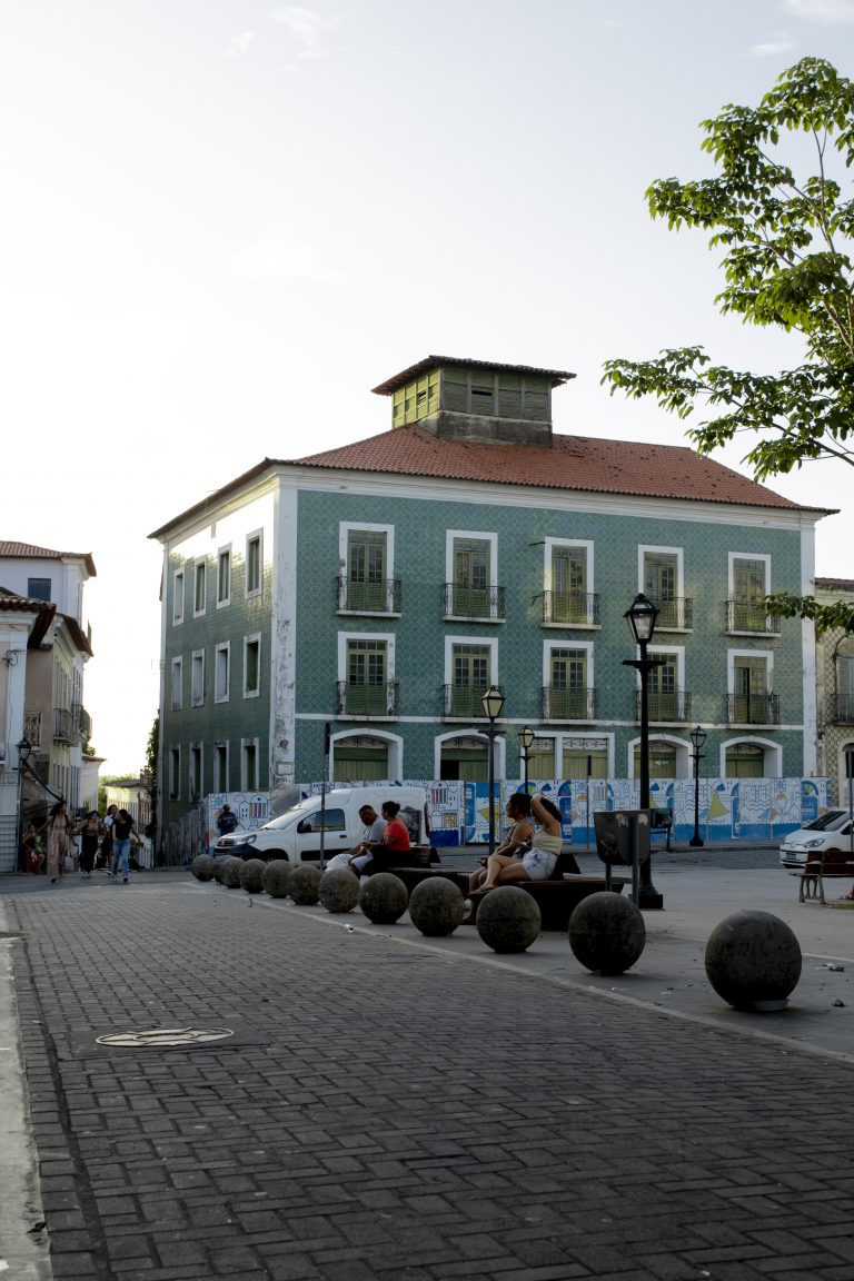 Algumas pessoas estão sentadas ou conversando perto das esferas. Há também postes de luz estilo colonial e uma árvore com folhas verdes no lado direito. O céu está claro, indicando que a foto foi tirada durante o dia.A imagem mostra um prédio histórico de três andares, pintado de verde-azulado, com janelas brancas e sacadas com grades pretas. O telhado é vermelho e tem uma estrutura quadrada no topo.
