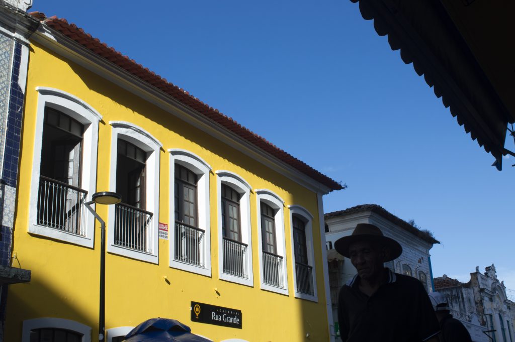 Fotografia de uma rua com arquitetura colonial, destacando-se um edifício amarelo vibrante de dois andares com janelas altas e sacadas com grades de ferro pintadas de branco. As molduras das janelas também são brancas, contrastando com o tom vivo da fachada. O prédio tem telhado de cerâmica vermelha e uma placa preta com letras brancas na parte inferior onde se lê: “Galeria Rua Grande”. À direita, parcialmente em silhueta por conta da sombra, aparece um homem idoso de pele escura, usando chapéu de aba larga e camisa preta com gola branca. Ao fundo, outros prédios antigos em tons mais neutros compõem a paisagem arquitetônica. O céu está completamente azul e sem nuvens, com iluminação forte do sol incidindo diretamente sobre os edifícios.