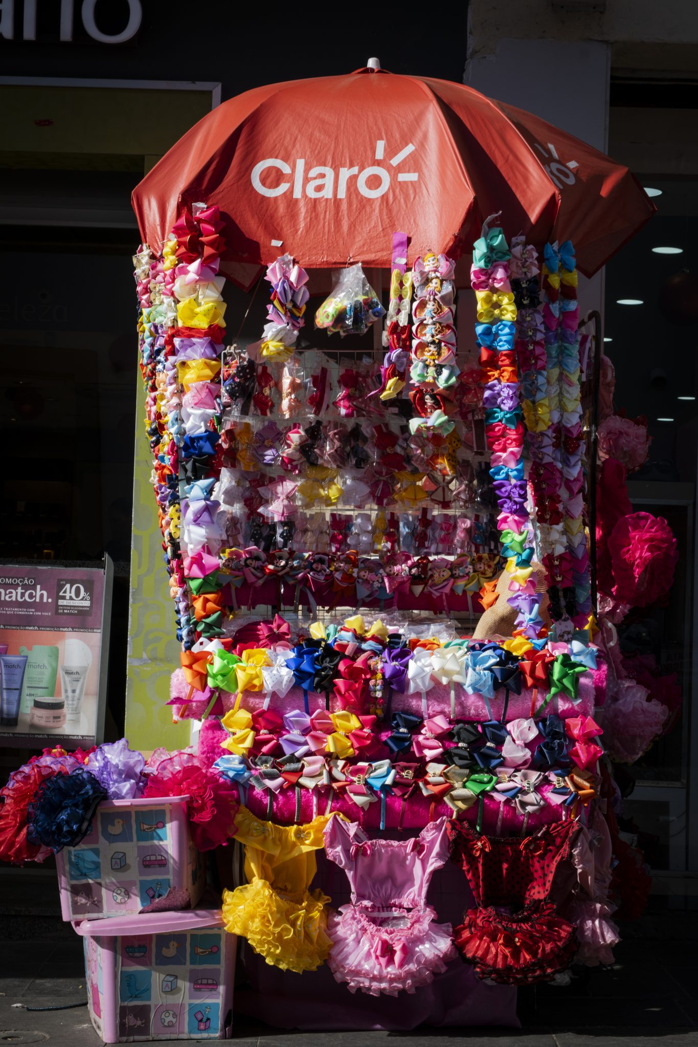 Em um ponto de venda de rua, um carrinho exibe uma grande variedade de acessórios coloridos, principalmente laços e fitas para cabelo. O carrinho é coberto por um grande guarda-sol vermelho com a marca “Claro” estampada no topo. Os laços estão organizados em fileiras, com cores vibrantes como rosa, amarelo, azul, verde e vermelho. Na parte inferior do carrinho, estão expostos pequenos vestidos infantis com babados e tecidos brilhantes, em cores chamativas como amarelo, rosa e vermelho com preto. Ao lado do carrinho, há caixas plásticas decoradas, contendo mais produtos, e um suporte com mais laços e adereços pendurados. Ao fundo, observa-se a fachada de uma loja iluminada e uma placa publicitária anunciando produtos de beleza com desconto.