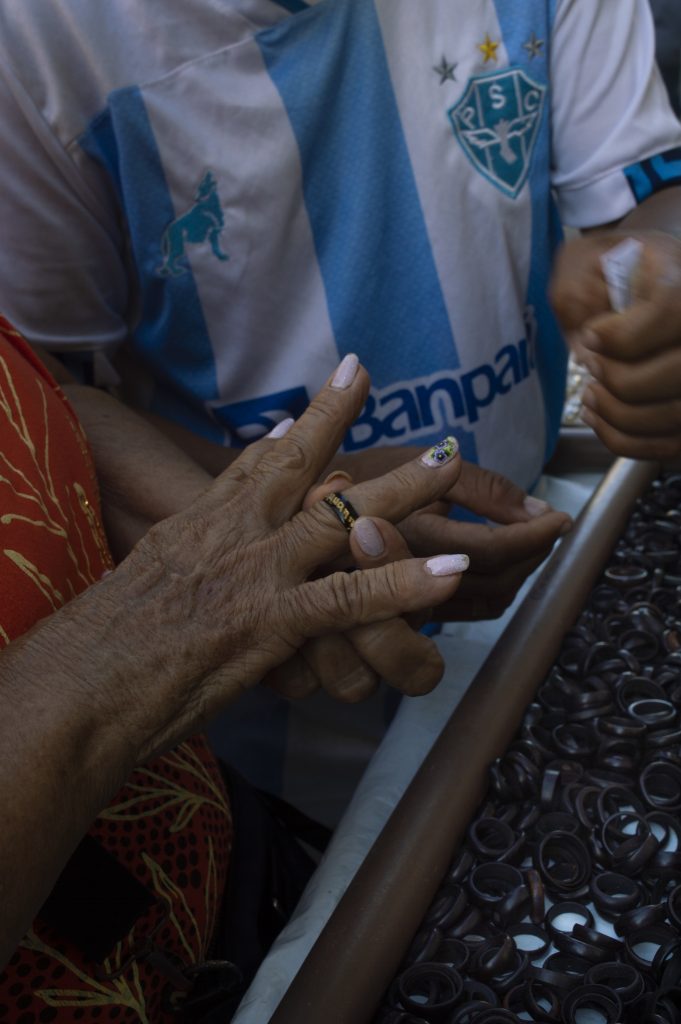 A imagem foca em um momento de interação entre mãos. Em primeiro plano, a mão de uma pessoa idosa, com pele enrugada e unhas cuidadas, uma delas decorada com desenho floral, experimenta um anel preto de tucum em um dos dedos. Ao fundo, parcialmente visível, está um homem com camisa listrada azul e branca do time Paysandu Sport Club, com logotipos e brasão em destaque. Sua mão também aparece, ajudando ou entregando o anel. Do lado direito, há uma bandeja repleta de anéis pretos organizados. A cena se passa provavelmente em uma feira ou banca de rua, sugerindo um momento simples de escolha e troca. A composição valoriza o gesto, o toque e o cotidiano.