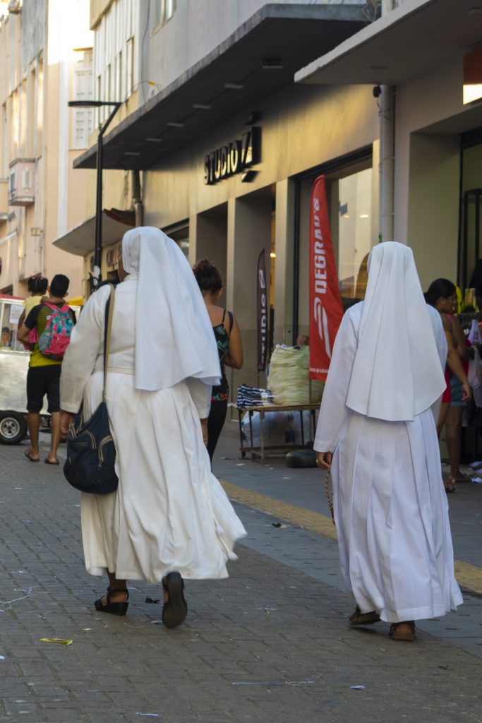 A imagem mostra uma rua de comércio popular, movimentada por pedestres. Em primeiro plano, duas freiras caminham lado a lado, vistas de costas. Elas vestem hábitos brancos longos com véus que cobrem a cabeça e parte das costas. A freira à esquerda carrega uma mochila preta pendurada no ombro; a da direita segura um terço marrom na mão. À frente delas, outras pessoas caminham, entre elas uma mulher com blusa preta estampada e alças finas. No lado direito da imagem, há barracas e expositores de roupas e produtos diversos. Uma loja com fachada moderna exibe a marca "Studio Z" e, ao lado, uma bandeira vertical vermelha com a palavra “DEGRAUS”. O chão é de piso intertravado, com uma faixa guia tátil amarela para pessoas com deficiência visual. O ambiente sugere fim de tarde, com luz suave e sombras alongadas projetadas na calçada.