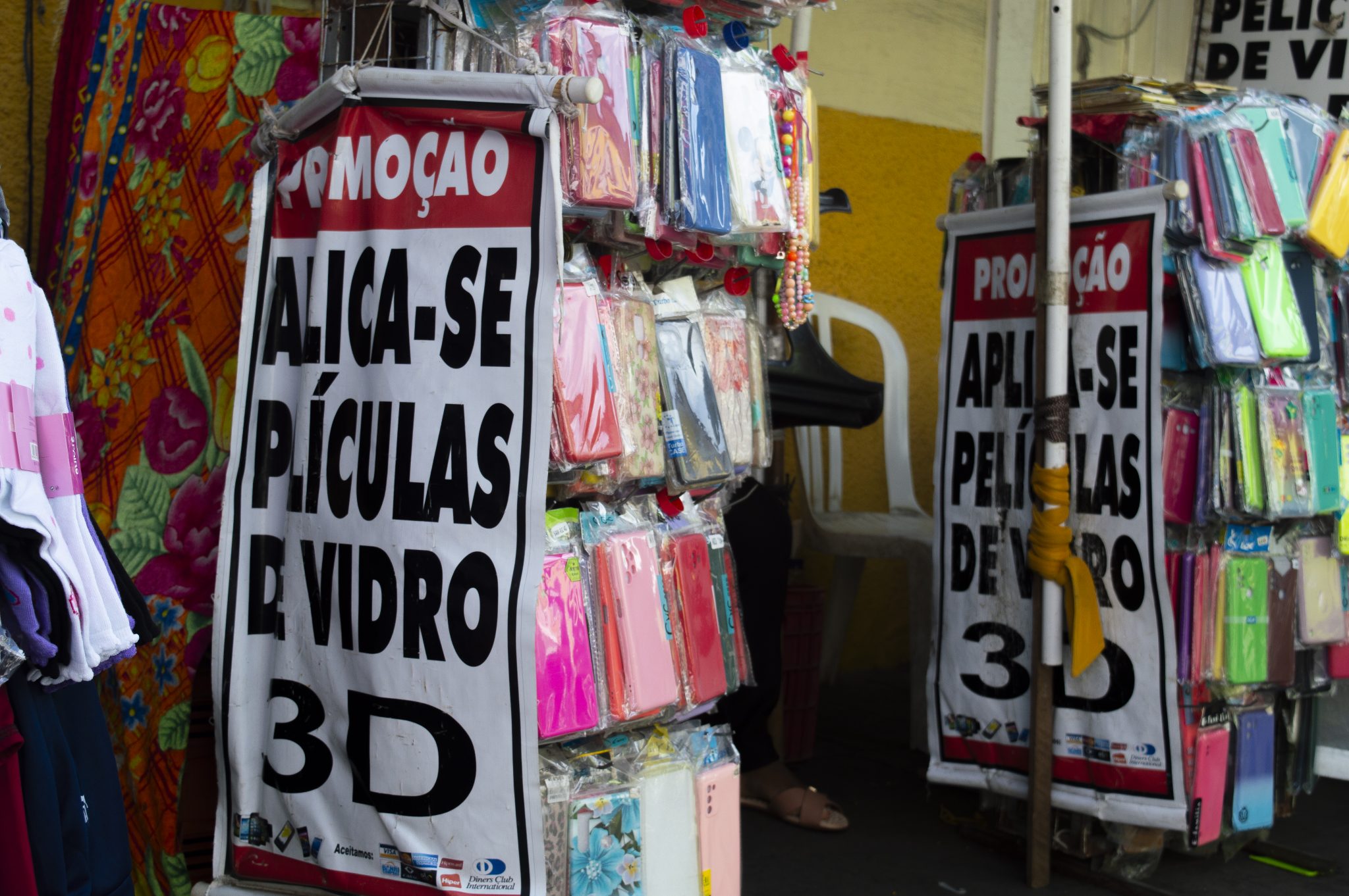 A fotografia retrata um cenário típico do comércio informal na Rua Grande, onde capinhas de celular estão expostas para venda. Os produtos, organizados sobre uma superfície simples, apresentam cores e detalhes variados que chamam a atenção. Ao fundo, observa-se a presença de construções com a identidade própria da rua, enquanto a movimentação de pessoas reforça o ambiente comercial e popular. A iluminação natural ressalta as texturas dos itens expostos, contribuindo para a compreensão do espaço e da cultura do comércio local.