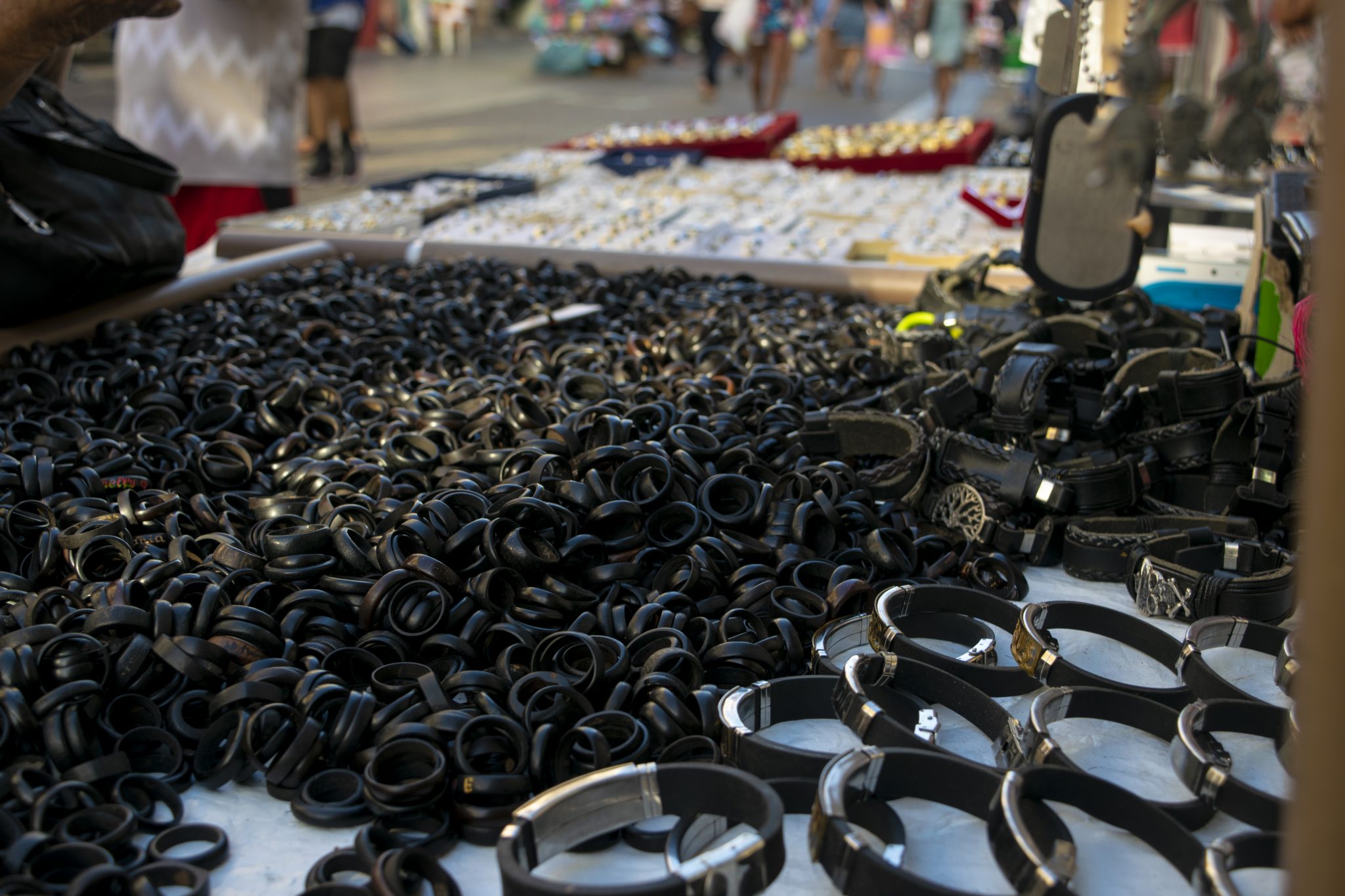 Em primeiro plano, há uma grande quantidade de anéis de coco na cor preta dispostos sobre uma superfície plana, formando um aglomerado compacto. Ao lado, pulseiras também pretas estão organizadas em fileiras, exibindo detalhes metálicos e fechos diferenciados. O cenário sugere uma banca ou mesa de vendas ao ar livre. Ao fundo, parcialmente desfocados, aparecem outras bancas com itens de bijuteria ou acessórios, além de pessoas circulando. A luz do final da tarde incide sobre os anéis e pulseiras, criando pequenos reflexos nos materiais e conferindo um tom dourado suave às bordas dos objetos.