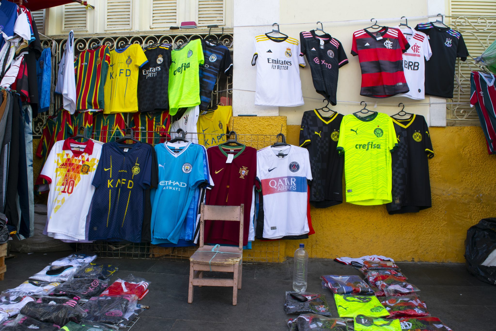 Expositor de rua com diversas camisetas de times de futebol penduradas em grades e cabides fixados a uma parede amarela. As camisetas, majoritariamente réplicas, representam clubes nacionais e internacionais como Flamengo, Palmeiras, Real Madrid, PSG, Borussia Dortmund, Manchester City, entre outros. Há modelos com diferentes cores e detalhes, como verde-limão fluorescente, vermelho e preto, azul claro e branco. Na frente do expositor, camisetas adicionais estão dobradas e embaladas em sacos plásticos sobre o chão de cimento. Ao centro da imagem, uma cadeira de madeira pequena e gasta está posicionada entre as roupas. Uma garrafa plástica reutilizada está ao lado da cadeira. À esquerda, algumas peças de roupa pendem de cabides improvisados.