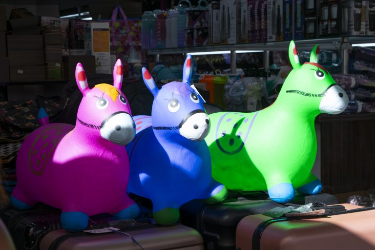 A imagem mostra três brinquedos infláveis em formato de cavalinhos dispostos lado a lado, expostos em uma loja na Rua Grande. Eles possuem cores vibrantes: o primeiro é rosa, o segundo azul e o terceiro verde. Cada cavalinho tem olhos grandes e expressivos, orelhas pontudas e focinho arredondado. Detalhes como manchas ou desenhos estão presentes nos corpos dos brinquedos, acrescentando um toque decorativo. Ao fundo, é possível ver prateleiras com diversos produtos, um ambiente comercial movimentado, típico da Rua Grande, em São Luís. A iluminação do local destaca as cores chamativas dos brinquedos.