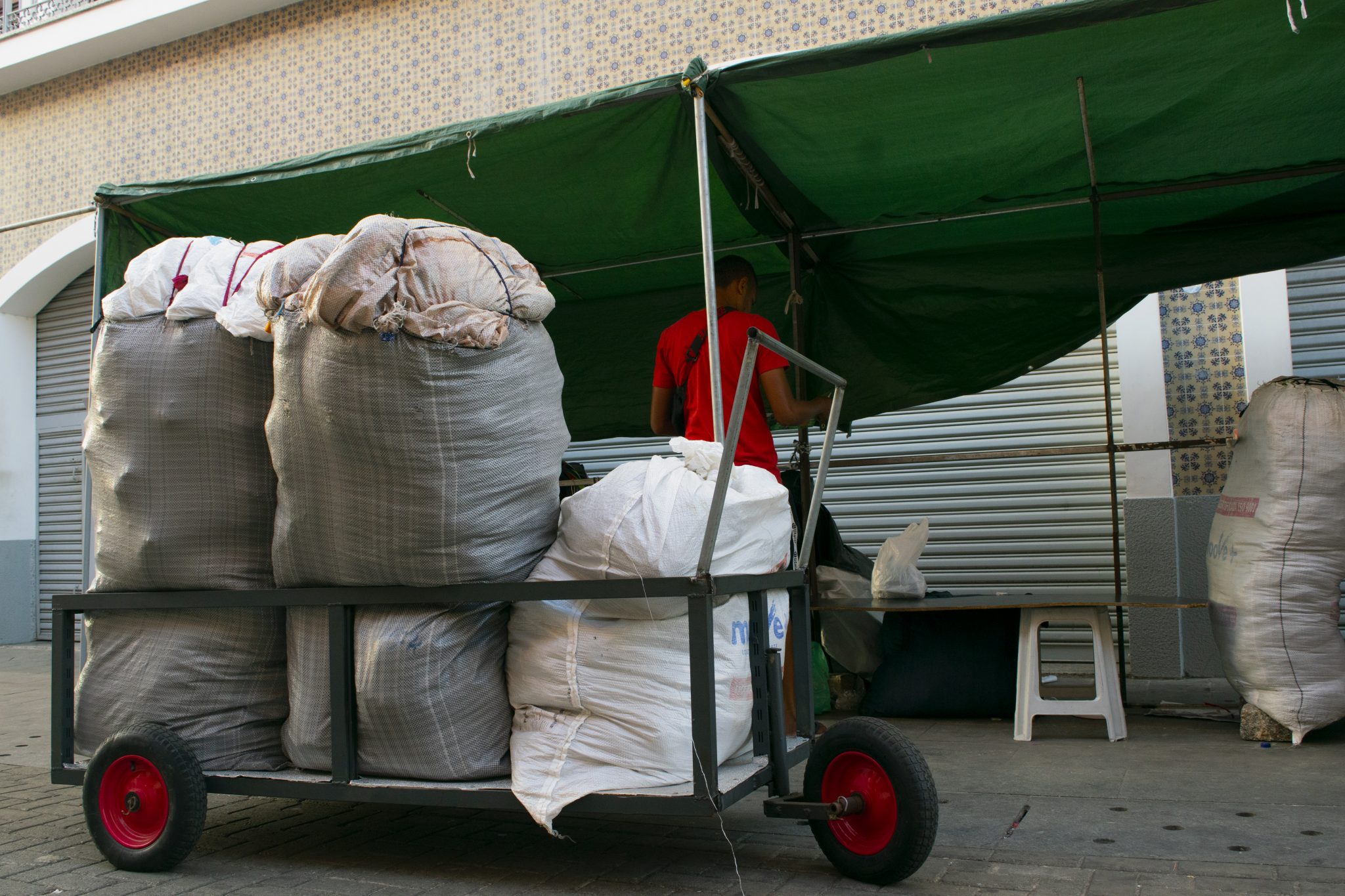 Cena de rua retratando um carrinho de carga metálico com rodas vermelhas, carregado com grandes sacos volumosos de ráfia, amarrados e repletos de mercadorias. Os sacos ocupam todo o espaço do carrinho e estão dispostos em pé, comprimidos uns contra os outros. Ao fundo, um homem de camiseta vermelha, de costas, está parcialmente coberto por uma lona verde presa em estrutura metálica típica cobertura de barraca de feira. O ambiente sugere o início de montagem de uma banca de comércio ambulante em frente a uma loja fechada com porta de metal enrolável. No canto direito da imagem, outro grande saco está no chão ao lado da barraca, enquanto uma mesa simples com uma sacola plástica e um banquinho branco completam a cena. O piso é de paralelepípedos e o prédio ao fundo tem azulejos decorativos azul e branco.