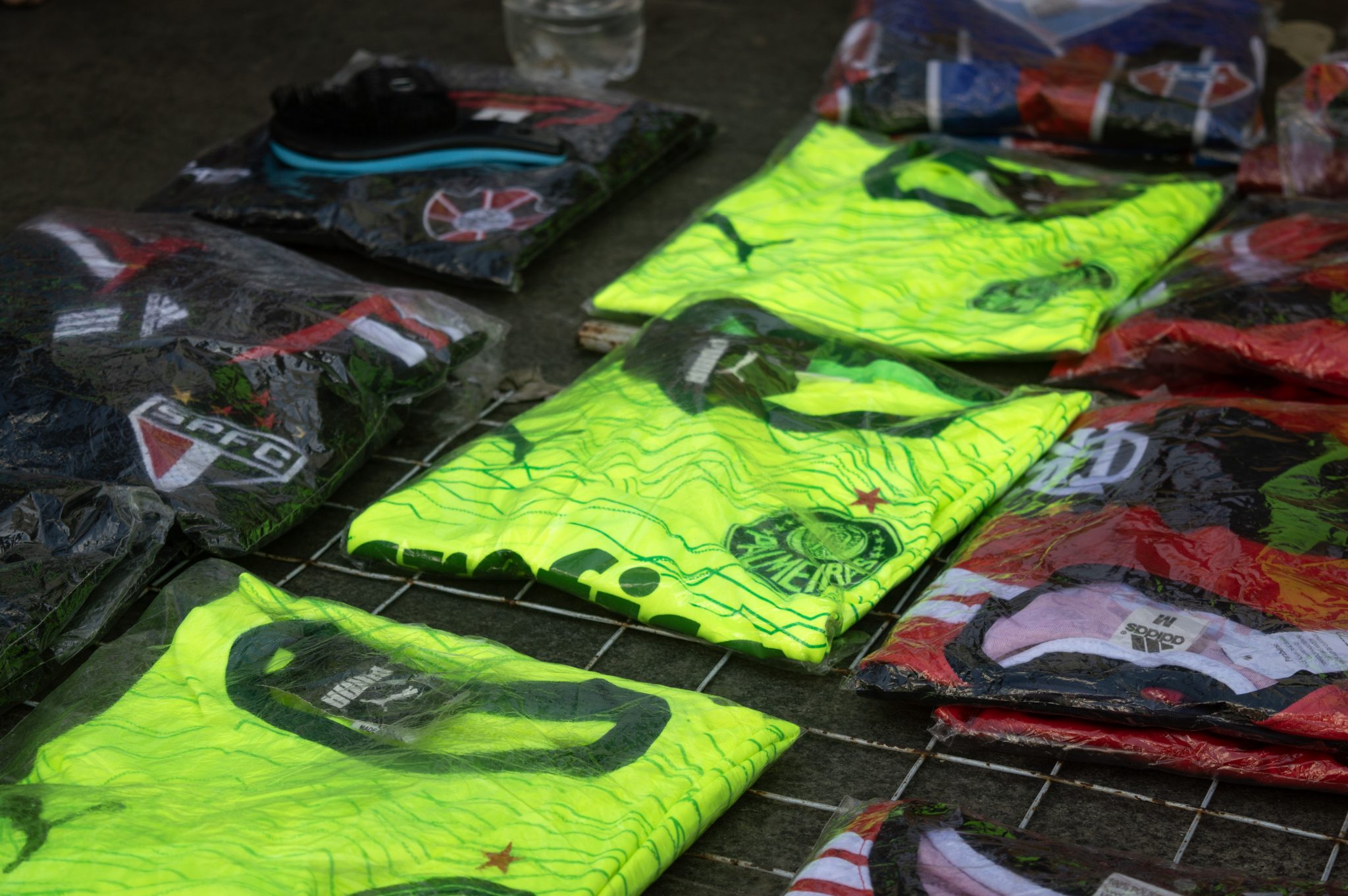 A imagem mostra camisas de times de futebol dispostas sobre uma grade no chão, em uma rua. As peças estão embaladas em plástico transparente, e algumas estão parcialmente abertas para exibir os detalhes. Em destaque, há camisas do Palmeiras com um design verde fluorescente e linhas onduladas, além do escudo do clube e uma estrela vermelha acima. Ao redor, há camisas de outros times, como Flamengo (predominantemente vermelho e preto), São Paulo (com as tradicionais listras tricolores), Moto Clube e Maranhão Atlético Clube, o MAC. O ambiente sugere um cenário de venda informal, característica da Rua Grande, com o objetivo de atrair clientes que passam pela rua.