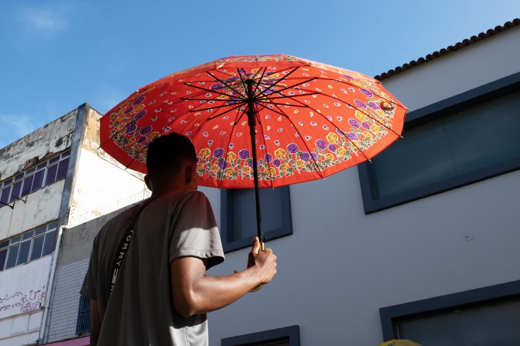 A fotografia mostra um vendedor de costas, em pé, segurando um guarda-chuva grande e colorido para se proteger do sol. O guarda-chuva é vermelho com detalhes florais em tons vibrantes, como amarelo, roxo e verde, que contrastam com o céu azul claro ao fundo. O homem veste uma camiseta cinza e está na Rua Grande, cercado por prédios. À esquerda, há um edifício antigo com pintura desgastada, enquanto à direita aparece parte de uma construção mais moderna com janelas de vidro e molduras escuras. A luz do sol é intensa, projetando sombras nítidas.