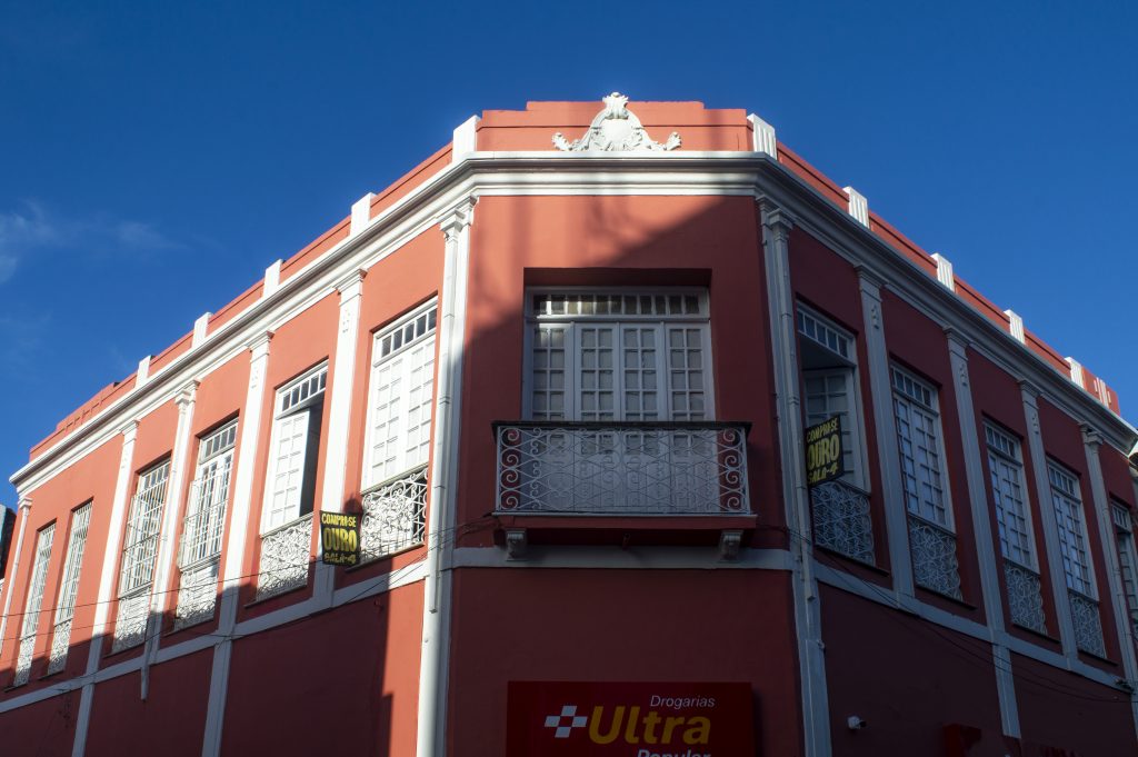 A imagem mostra a fachada de um prédio histórico de esquina, pintado em vermelho vibrante com detalhes brancos. O edifício possui dois andares e janelas altas com esquadrias e gradis brancos, todas simetricamente distribuídas. Em destaque, uma varanda com grade de ferro trabalhado sob uma das janelas centrais. No topo da construção, um frontão decorativo branco em estilo clássico enfeita o canto superior do prédio. Uma sombra projetada de outro edifício encobre parte da fachada, criando um contraste entre luz e escuridão. Na base do prédio, a placa de uma farmácia “Drogarias Ultra Popular” aparece em vermelho e amarelo. Cartazes nas janelas anunciam a compra de ouro. O céu está completamente azul, limpo, reforçando o clima ensolarado do dia. A arquitetura e a composição transmitem um ar de preservação cultural e vida urbana ativa.