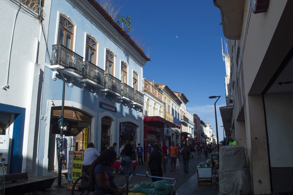 A imagem mostra uma movimentada rua de comércio popular em um centro histórico. À esquerda, um casarão azul claro com janelas altas emolduradas de branco e sacadas de ferro trabalhado chama a atenção. Abaixo, uma loja com a placa “CASAS BAHIA” funciona no térreo do prédio. Ao fundo, outros prédios antigos, coloridos e com arquitetura similar, formam uma linha contínua até o horizonte. Do lado direito da rua, barraquinhas, carrinhos de mercadorias e caixas cobertas compõem o cenário de vendedores ambulantes. Diversas pessoas caminham, algumas com sacolas, outras conversando ou observando vitrines. O céu está azul intenso, com a lua visível mesmo durante o dia. A iluminação natural e as sombras suaves indicam o fim da tarde. A cena retrata o cotidiano vibrante e popular de uma rua central.