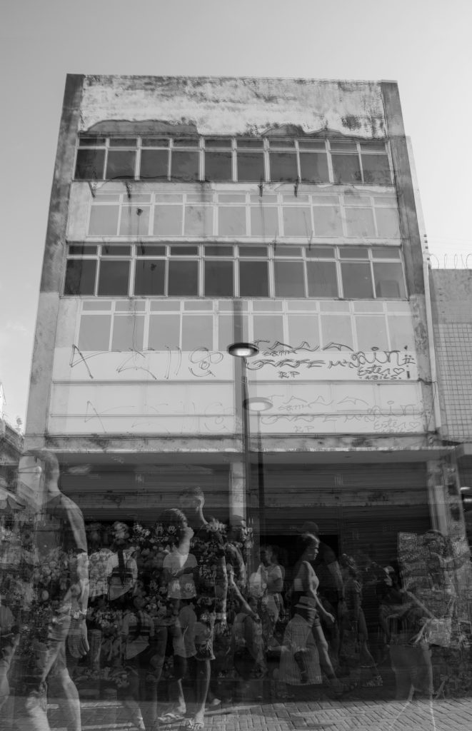 A imagem em preto e branco retrata uma cena urbana onde um edifício de quatro andares domina a composição. Sua fachada apresenta sinais evidentes de desgaste, com manchas escuras e descascamento na parte superior, além de janelas de vidro emolduradas por estruturas metálicas. Grafites e rabiscos cobrem parte da parede no nível térreo, trazendo um aspecto de abandono e resistência ao espaço arquitetônico. Sobrepondo-se ao prédio, um fluxo de pessoas caminha pela calçada, criando um efeito visual fantasmagórico. As silhuetas translúcidas sugerem um ritmo contínuo e dinâmico do comércio local, evocando a efemeridade do movimento urbano. Algumas figuras destacam-se mais nitidamente: um homem de camiseta clara e bermuda caminha à esquerda, enquanto uma mulher de vestido estampado avança pelo centro da imagem. Entre os transeuntes, formas sobrepostas revelam elementos comerciais, como balões e produtos expostos, insinuando a presença de barracas e vendedores ambulantes. Ao centro, um poste de iluminação se ergue, cortando verticalmente a composição. Sua luz não está acesa, mas sua presença contribui para a sensação de profundidade da cena. A textura do chão de paralelepípedos é visível em meio às camadas de imagens, reforçando a identidade histórica e comercial da área. A fusão entre o prédio estático e o movimento incessante das pessoas cria um contraste simbólico entre a permanência e a transitoriedade da vida urbana.