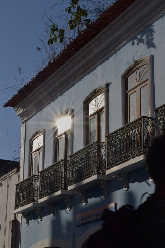 m varandas com grades de ferro trabalhadas em detalhes ornamentais. Um forte reflexo do sol incide sobre uma das janelas, criando um brilho intenso em forma de estrela que se espalha em feixes luminosos. No topo do prédio, o telhado de telhas cerâmicas se destaca, e pequenas plantas crescem entre as frestas e na calha, sugerindo ação do tempo e ausência de manutenção. A sombra de outro edifício cobre parte da fachada azul e da placa comercial “CASAS BAHIA”, na base da construção. No canto inferior direito da imagem, aparece parcialmente uma silhueta escura de uma pessoa, desfocada, contribuindo com uma sensação de movimento e vida urbana. O céu ao fundo está limpo, azul profundo.