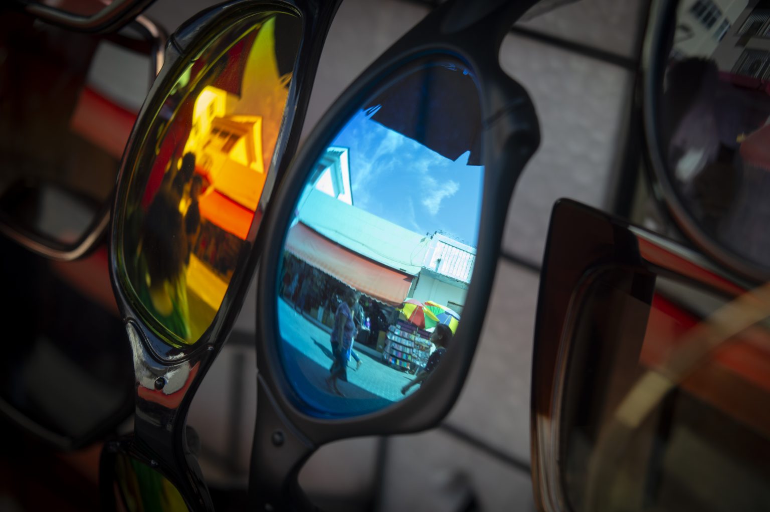 Transeuntes no reflexo de lentes de óculos à venda por camelôs na Rua Grande, em São Luís. A imagem mostra de perto três pares de óculos com lentes espelhadas, presos a uma grade de exposição. Cada lente reflete uma cena da rua: a lente amarela mostra pessoas caminhando diante de fachadas em tons quentes; a lente azul reflete o céu claro, barracas de lona laranja e uma movimentada calçada; a lente à direita, com tonalidade escura, também mostra partes do ambiente urbano. O fundo está desfocado, dando destaque aos reflexos nas lentes. A imagem é vibrante e possui contrastes de cores.