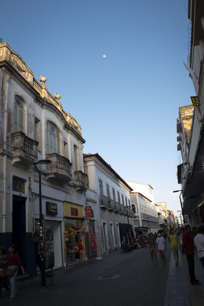 A imagem retrata uma cena urbana da Rua Grande, em São Luís do Maranhão, durante o entardecer. O céu azul domina a parte superior da fotografia, com a Lua visível ao fundo, desfocada e distante, adicionando um toque celestial à composição. À esquerda, destacam-se edificações históricas de estilo colonial, com fachadas ornamentadas e varandas de ferro trabalhado. As janelas altas e os detalhes arquitetônicos remetem ao período de influência portuguesa na região. Do lado direito, há construções mais simples, mas ainda preservando traços históricos. A rua está movimentada, com pessoas caminhando em ambas as direções. Algumas estão próximas às lojas que ocupam o térreo dos edifícios, enquanto outras seguem em pequenos grupos. As vitrines exibem produtos variados, incluindo roupas e acessórios, sugerindo um espaço comercial ativo e popular.