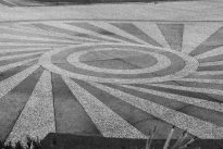Fotografia retangular de um desenho no chão, composto por formas geométricas. O chão tem tons de cinza. No centro, um círculo maior, rodeado por linhas que se expandem.