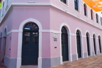 Fotografia horizontal ao dia de um casarão rosa com seis janelas ao topo e seis portas verdes semicirculares com bordas brancas,no canto superior direito bandeirinhas laranjas.