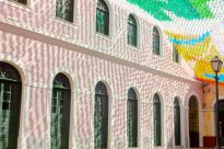 Fotografia retangular da fachada de um prédio cor de rosa com janelas e portas verdes semi-circulares. No canto superior direito da imagem, bandeirinhas de São João com sombras projetadas na parede.