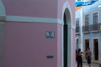 Fotografia horizontal ao dia de esquina de casarão cor de rosa com placa metálica “Rua da Estrela” no centro,ao lado porta verde semicircular com borda branca,ao fundo lateral parte de um casarão azul.