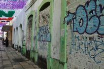 Fotografia vertical, uma calçada antiga rodeada por prédios baixos e degradados. O muro à direita possui um graffiti em tons de azul e verde. no canto superior, à esquerda, bandeiras coloridas.