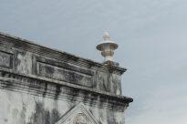 Fotografia horizontal ao dia de um beiral de construção antiga,com peça decorativa de um vaso ornamental branco sobre alvenaria escurecida pelo tempo,ao fundo um céu azul com nuvens.