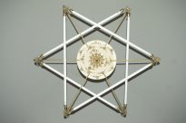 Fotografia horizontal de luminária metálica em forma de estrela de seis pontas,com detalhes metálicos em cada ponta,no centro da luminária, uma estrutura redonda,duas sobreposições e detalhes dourados em formato de mandala.