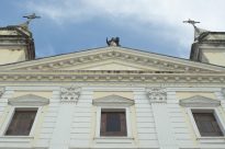 Fotografia horizontal ao dia,vista de baixo para cima da parte superior triangular da igreja da Sé,com escultura ao topo de Santa Vitória e duas cruzes nas torres laterais,abaixo duas colunas brancas verticais e três janelas de madeira no centro.Ao fundo céu azul com nuvens.