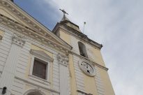 Fotografia horizontal ao dia de parte lateral da igreja da Sé amarela e branca,com cruz metálica no topo,uma janela retangular vertical, ao lado relógio circular quebrado e ao fundo um céu azul com nuvens.