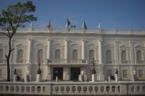 Fotografia horizontal ao dia da fachada branca do Palácio dos Leões,sacadas com três bandeiras pequenas hasteadas no topo,à esquerda bandeira do Maranhão,ao centro bandeira do Brasil,à direita bandeira verde,abaixo nove janelas simétricas semicirculares,e ao fundo,céu azul com nuvens