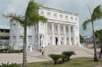 Fotografia horizontal ao dia, da fachada branca da Biblioteca Benedito Leite, com seis colunas altas e circulares na entrada e sete janelas alinhadas no andar superior escadaria à frente e coqueiro em primeiro plano ao lado inferior esquerdo da cena, fundo céu azul com nuvens.
