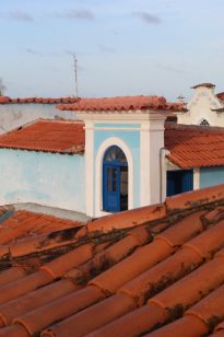 Fotografia horizontal ao dia de parte de um casarão azul com detalhes brancos,janela semicircular azul no centro e telhados cerâmicos,ao fundo,céu azul com nuvens.