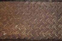 Fotografia retangular de uma chapa de ferro enferrujada em tons de marrom. A textura é marcada por relevos geométricos irregulares e repetidos.