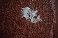 Fotografia retangular de uma parede vermelha com textura irregular chapiscada. No centro, uma pequena mancha branca.