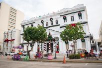 Fotografia horizontal em uma visão ampla. Em contraste com o céu azul, construção histórica branca decorada com festões verdes, laços vermelhos e guirlandas sobre as janelas. À frente uma árvore de Natal artificial com base listrada em branco e vermelho, cercada por duas árvores naturais de folhagem escura. Pessoas estão sentadas em bancos próximos, um carrinho de comida roxo e branco à esquerda.