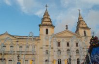 Fotografia horizontal da fachada de uma igreja histórica em tons claros, com duas torres altas e cruz no topo de cada uma. Na frente do prédio, jatos de água de uma fonte arqueiam para o alto, criando linhas translúcidas que se destacam contra o céu azul. À direita, uma decoração natalina, com enfeites coloridos e luzes.