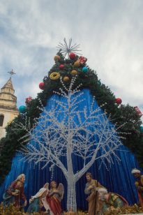 Fotografia vertical, com grande decoração natalina.Acima, uma árvore decorativa branca e um arco de folhagem verde com bolas vermelhas, douradas e azuis, além de pequenos presentes, finalizada por uma estrela metálica. No centro, um painel azul brilhante serve de fundo para um presépio com figuras com anjos, pastores e reis magos. À esquerda, parte de uma torre antiga de igreja com uma cruz.
