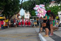 Fotografia horizontal, ao fundo o céu azul, em um espaço urbano com árvores, barracas de comida, pessoas observando e veículos ao fundo. No centro cinco músicos vestidos como duendes natalinos, com meias listradas, sapatos pontudos e gorros utilizando instrumentos de sopro.