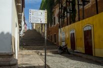 Fotografia horizontal ao dia de placa de sinalização turística,ao lado um casarão antigo em ruínas com folhagens verdes cobrindo canto lateral esquerdo,ao fundo escadaria de pedra e céu azul.