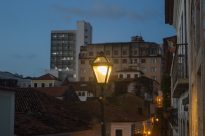 Fotografia horizontal à noite,poste colonial iluminado,com prédios altos antigos ao fundo.